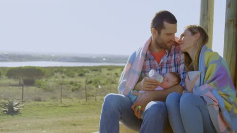 Front-view-of-mid-adult-caucasian-parents-feeding-their-baby-from-a-bottle-in-back-yard-of-home-4k