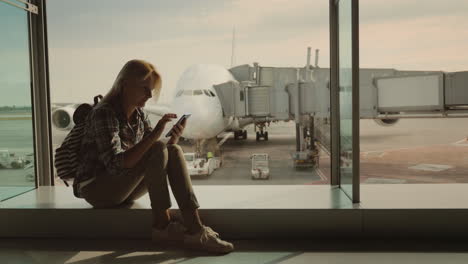 a female passenger sits on the windowsill in the airport terminal uses a smartphone against the back