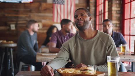 man watching game on screen in sports bar eating burger and fries and drinking beer