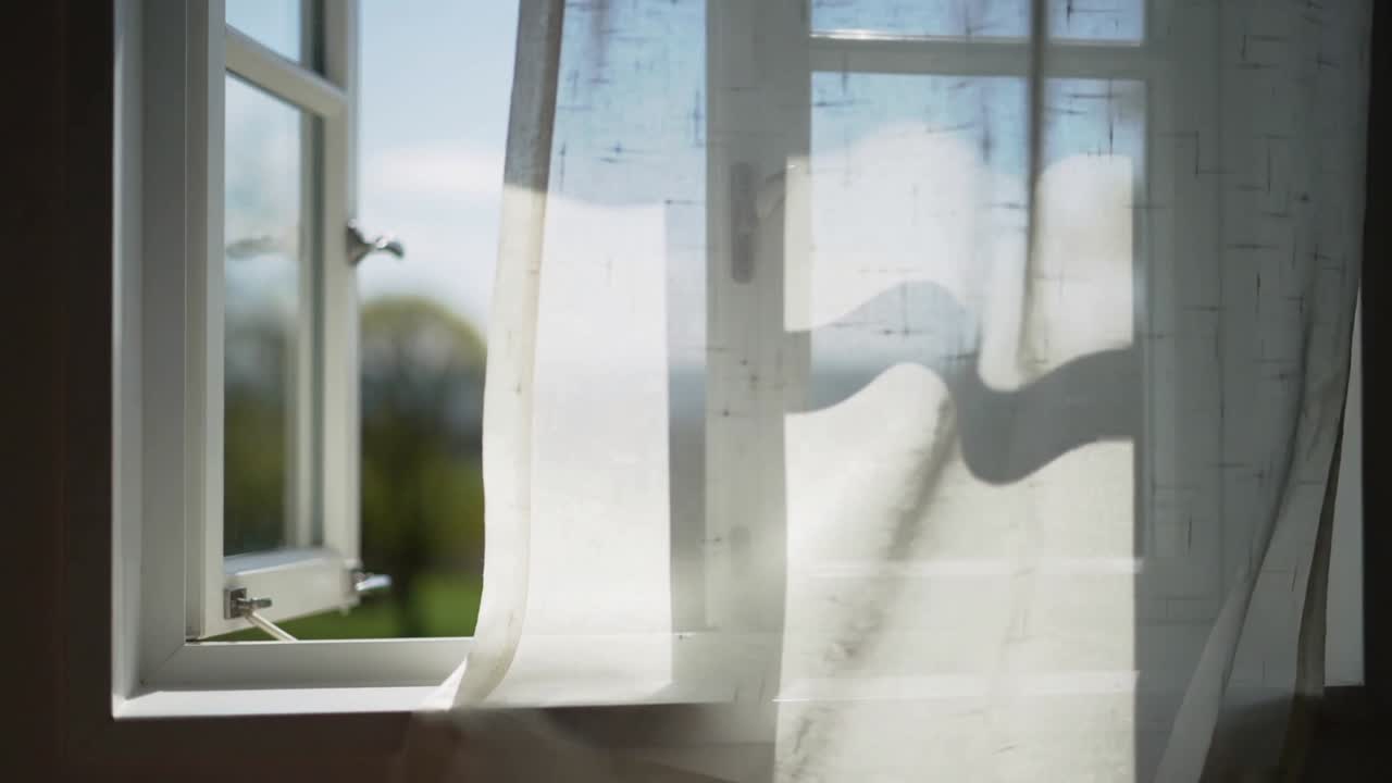 Ein wehender, transparenter Vorhang, der auf eine Brise reagiert, die durch ein links angeschlagenes Fenster im Haus weht und die Fenstersprossen sichtbar macht.