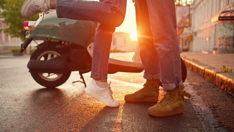 Close-up-shot-of-a-girl-standing-on-her-toes-to-hug-her-boyfriend-near-their-moped-on-the-street-during-dawn-in-summer.-Legs-of-a-guy-and-a-girl-in-denim-pants-standing-opposite-each-other-against-of-a-bright-sunny-dawn-in-summer