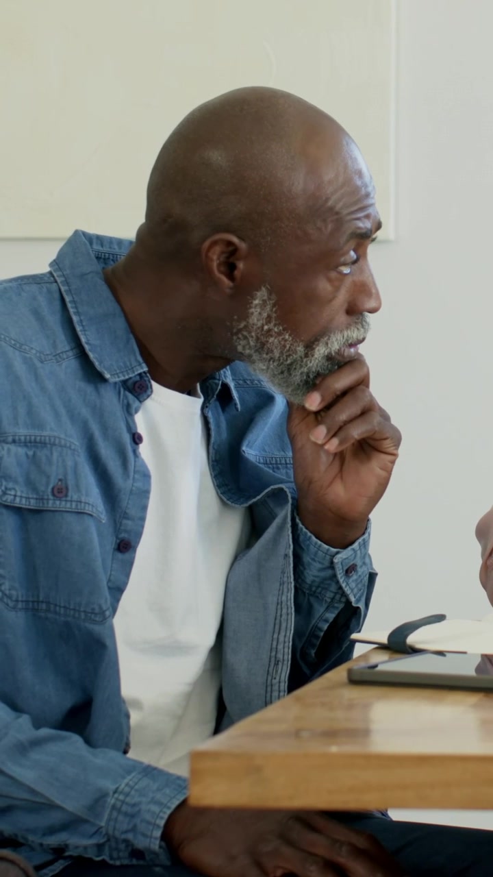 Vertical video: Listening man in denim leaning at home table while partner talking, pointing tablet