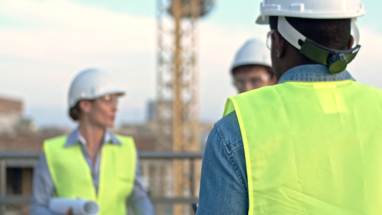 vista desde atrás del hombre afroamericano constructor en sombrero duro que viene a sus colegas caucásicos - hombre y mujer mientras hablan al aire libre en el sitio de construcción.