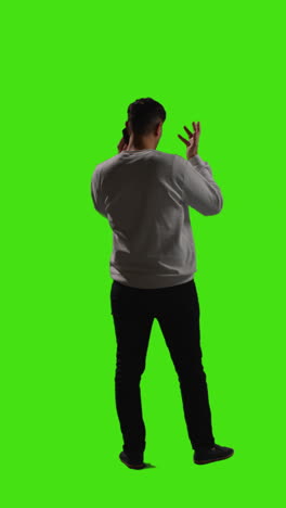 Vertical-Video-Full-Length-Rear-View-Shot-Of-Young-Man-Talking-On-Mobile-Phone-Standing-Against-Green-Screen-With-Low-Key-Lighting