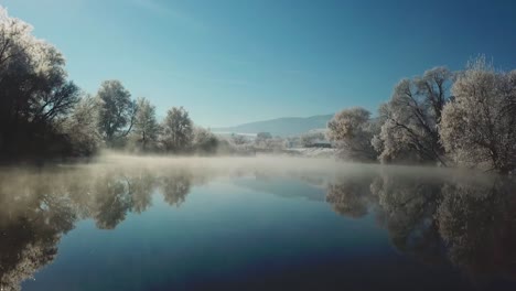 Eisnebel-Auf-Gefrorener-Seeoberfläche,-Ausgerichtet-Von-Schneebedeckten-Bäumen-Im-Kalten-Winter,-Dolly-In-Schuss