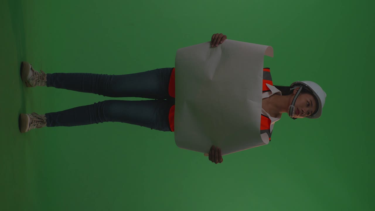 cuerpo lleno de ingeniera asiática con casco de seguridad mirando el plano en sus manos y mirando a su alrededor mientras está de pie en el estudio de fondo de pantalla verde, durante la puesta o el amanecer