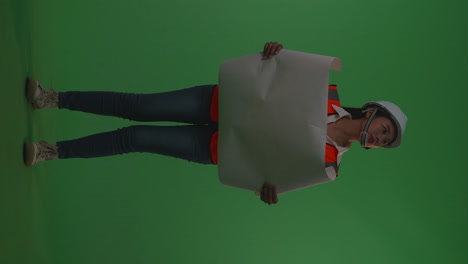full body of asian female engineer with safety helmet looking at blueprint in her hands and looking around while standing in the green screen background studio, during sunset or sunrise time