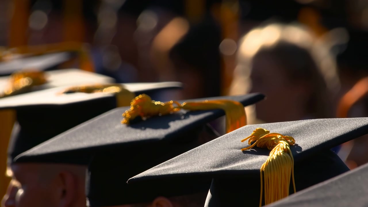 Lavt skråperspektiv av rader med svarte studenthatter med gullkvaster under varmt utendørs lys, som viser subtil bevegelse i kvastene, myk bokeh og en mild feiringstemning.