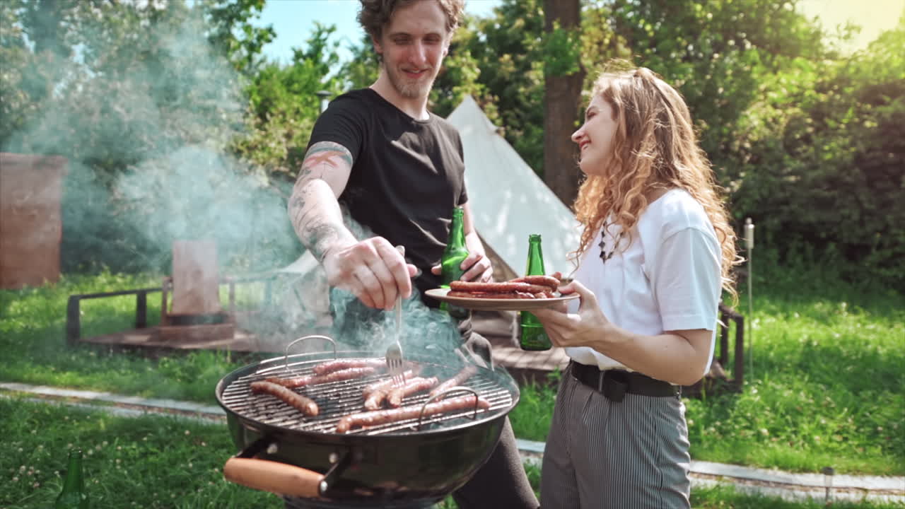 Young couple frying meat on the grill with beer and putting it on the plate. Greenery around. Glamping. Slow motion