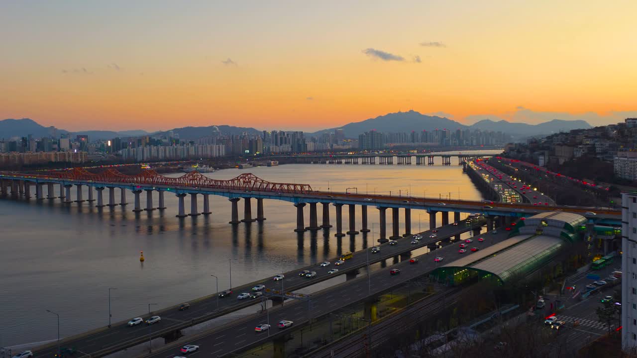 Inquadratura zoom del paesaggio urbano di Seoul al tramonto sul fiume Han, con il Ponte Donho, le luci incandescenti del traffico autostradale, le montagne e lo skyline della città.