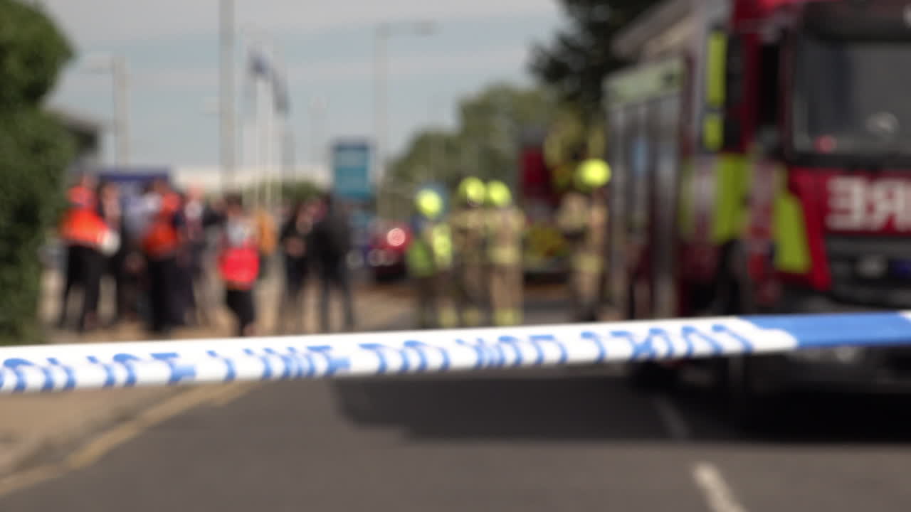 Police cordon tape flutters in the wind in front of firefighters, government representatives and fire engines at the scene of a tower block covered in flammable cladding that was destroyed by a fire.