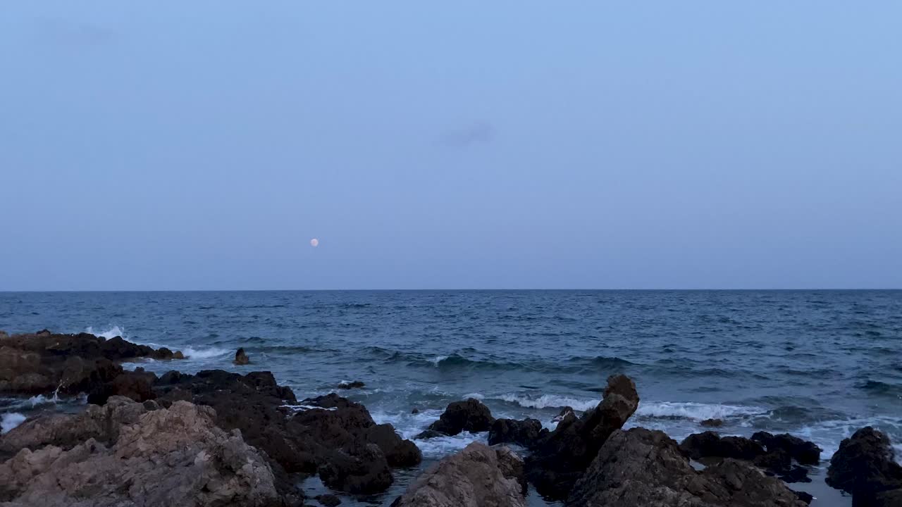 해가 지면 바위가 있는 해안선에 파도가 부히고, 뒤에 달이 뜨고 있다.