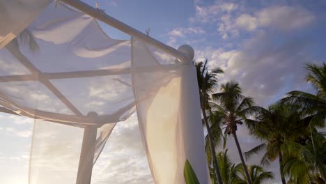 Detail-of-a-wedding-arch-made-of-light-fabric-set-against-a-tropical-sky-at-sunset