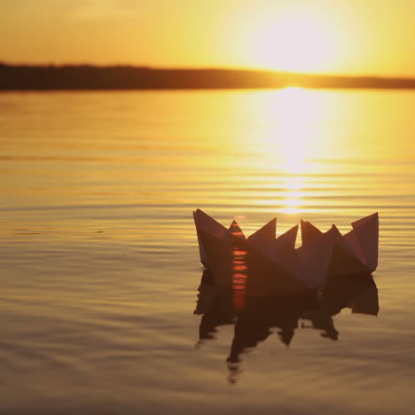 female's hand putting one paper boat on the water and it joins to other ones at sunset. new origami ship adding to the rest during beautiful sunset with the sun reflection in the lake