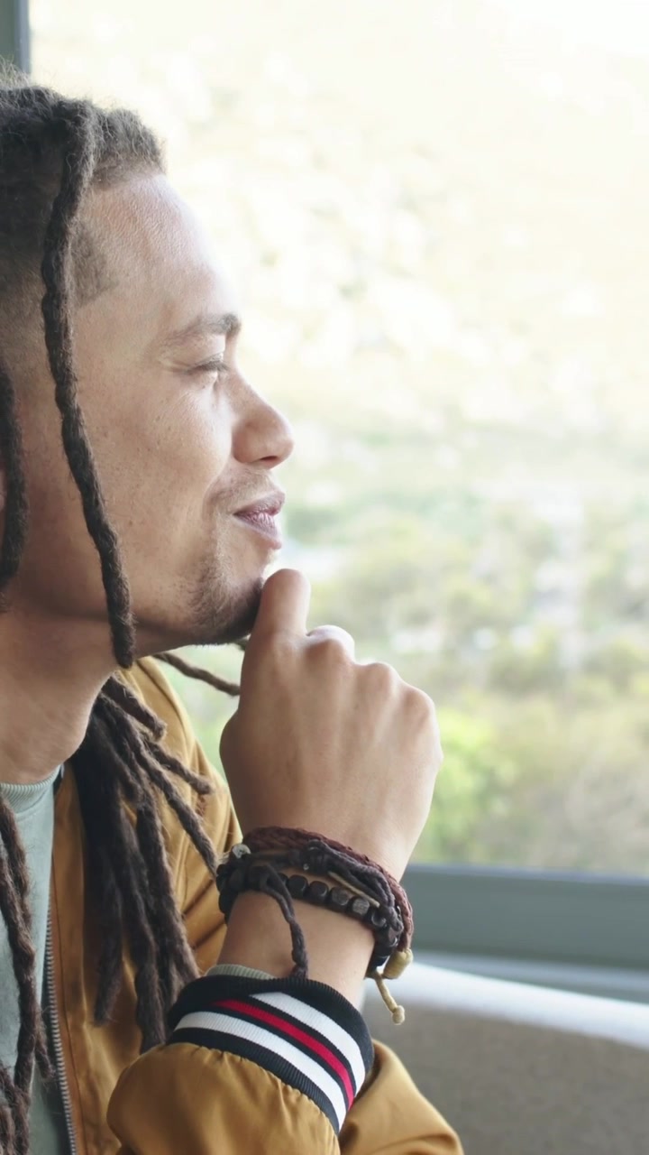 Video vertical: Hombre con chaqueta mostaza en el sofá, mirando por la ventana, tocándose la sien y reflexionando.