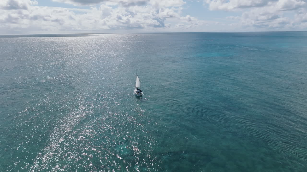 tomada aérea de un avión no tripulado de un yate de vela, mirando hacia el vasto y interminable horizonte sobre aguas sinuosas, salpicadas de nubes y olas contra un cielo azul luminoso en el mar caribe, méxico