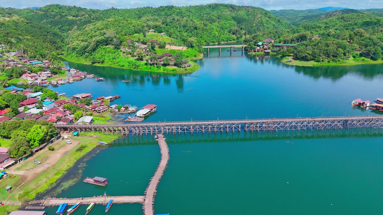 Drone shot rising above the Mon Bridge in Sangkhlaburi, Thailand. The long wooden structure crosses blue water, with floating houses, boats, and green hills in the background