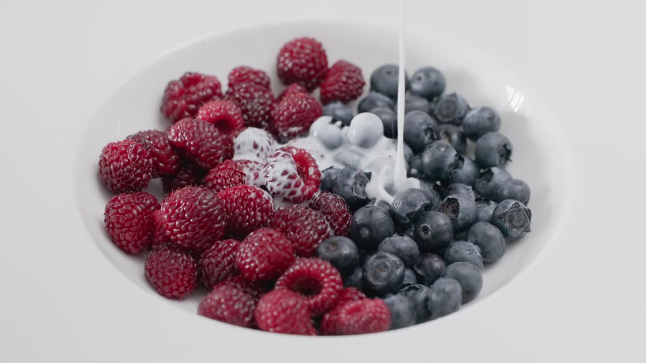 Close-up slow motion shot of creamy white yogurt being poured over a bowl of fresh, ripe organic raspberries and blueberries for a delicious and healthy antioxidant-rich breakfast or dessert.