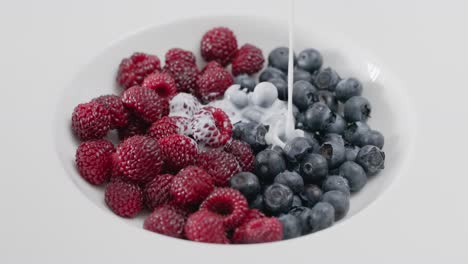 close-up slow motion shot of creamy white yogurt being poured over a bowl of fresh, ripe organic raspberries and blueberries for a delicious and healthy antioxidant-rich breakfast or dessert.