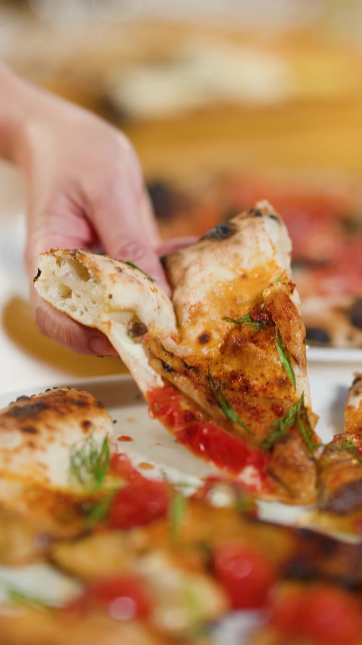 Una persona solleva una fetta di pizza cotta nel forno a legna con salsa di pomodoro e basilico fresco.