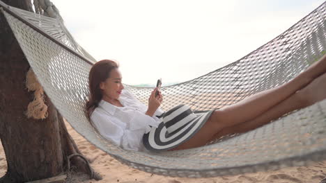 young asian woman laying on beach hamcock and looking at smartphone with smile