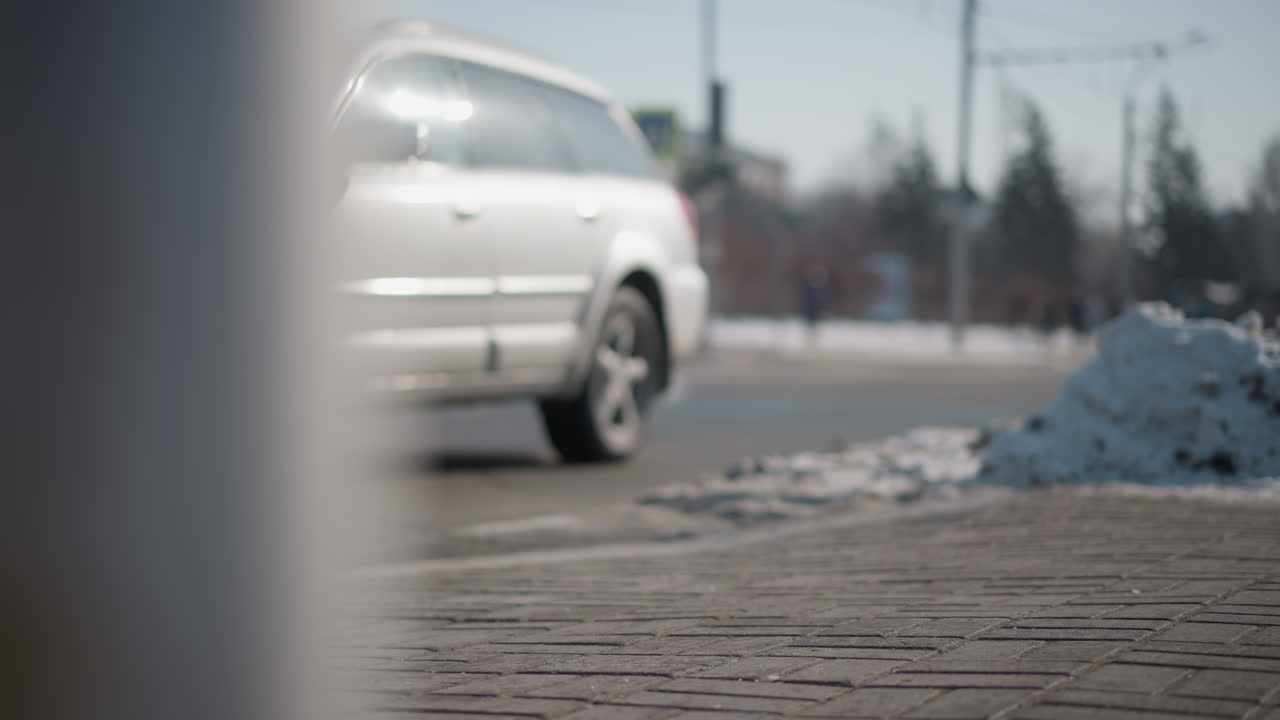 view past pole shows blur cars moving along busy winter road, brick pavement and curb in foreground, snow heap at corner catching sun, soft bokeh background, cold city commute vibe