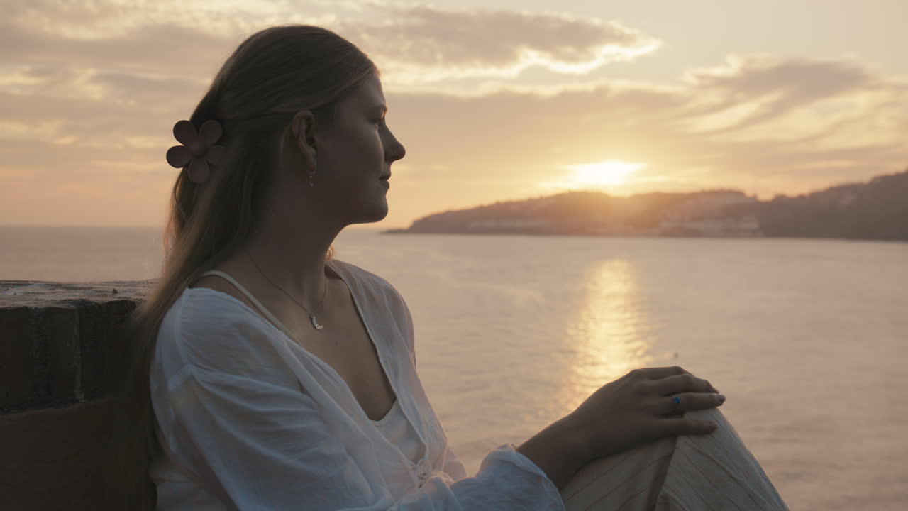 Medium side profile shot in golden hour light, showing a serene woman with an engagement ring, looking out over the ocean—evoking romance and reflection