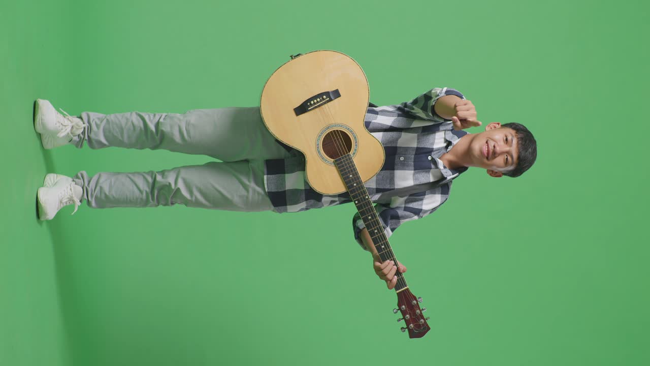 Adolescente asiatico a figura intera con la chitarra che fa un gesto onesto e sorride su sfondo a schermo verde