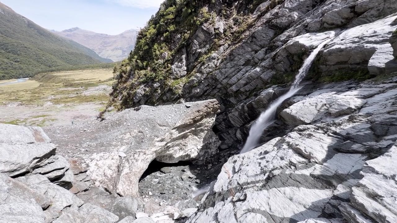 Cascata panoramica che scende su roccia di granito levigata sopra una vasta valle glaciale nel Parco Nazionale di Mount Aspiring. Catturata in una giornata di sole con aspro paesaggio alpino e natura incontaminata