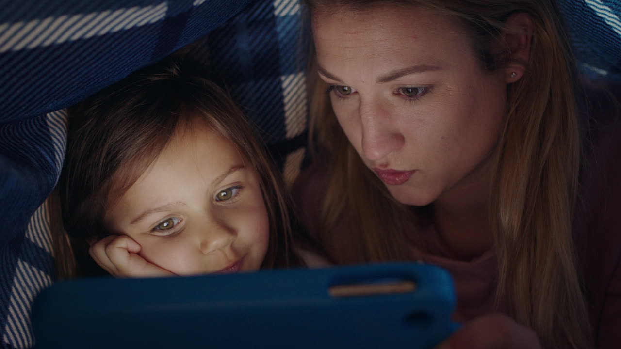 행복한 어머니와 딸은 침대  ⁇ 에서 태블릿 컴퓨터를 사용하고 터치 스크린 기술로 게임을 하며 휴식을 취하고 잠자리에 들기 전에 재미를 느끼고 있습니다.