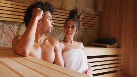 video of relaxed diverse couple wearing towels sitting and talking in sauna room at health spa