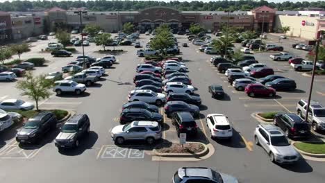 a crowded parking lot filled with cars of different sizes during midday. customers navigate through the area while storefronts line the background, creating a bustling shopping atmosphere.