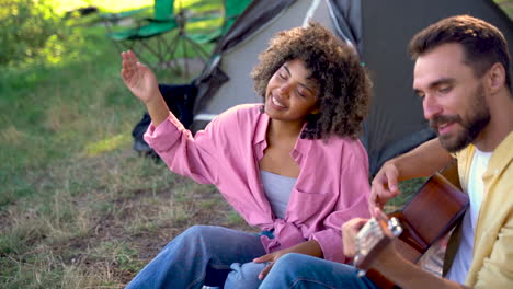 happy hiker couple camping in the forest. young black woman dances and sings, caucasian man plays guitar.