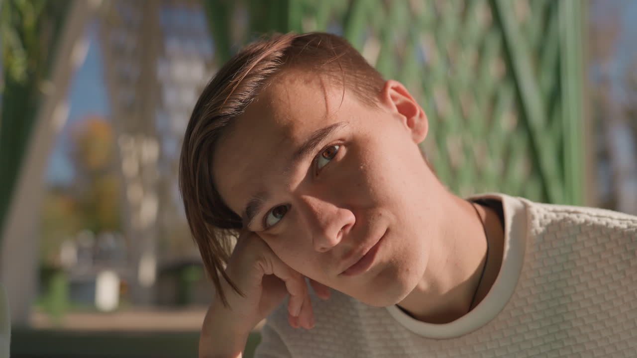 Pensive young white man outdoors, student leaning on hand, soft autumn light on face against green lattice backdrop, closeup portrait with introspective expression, cinematic shallow depth, quietly