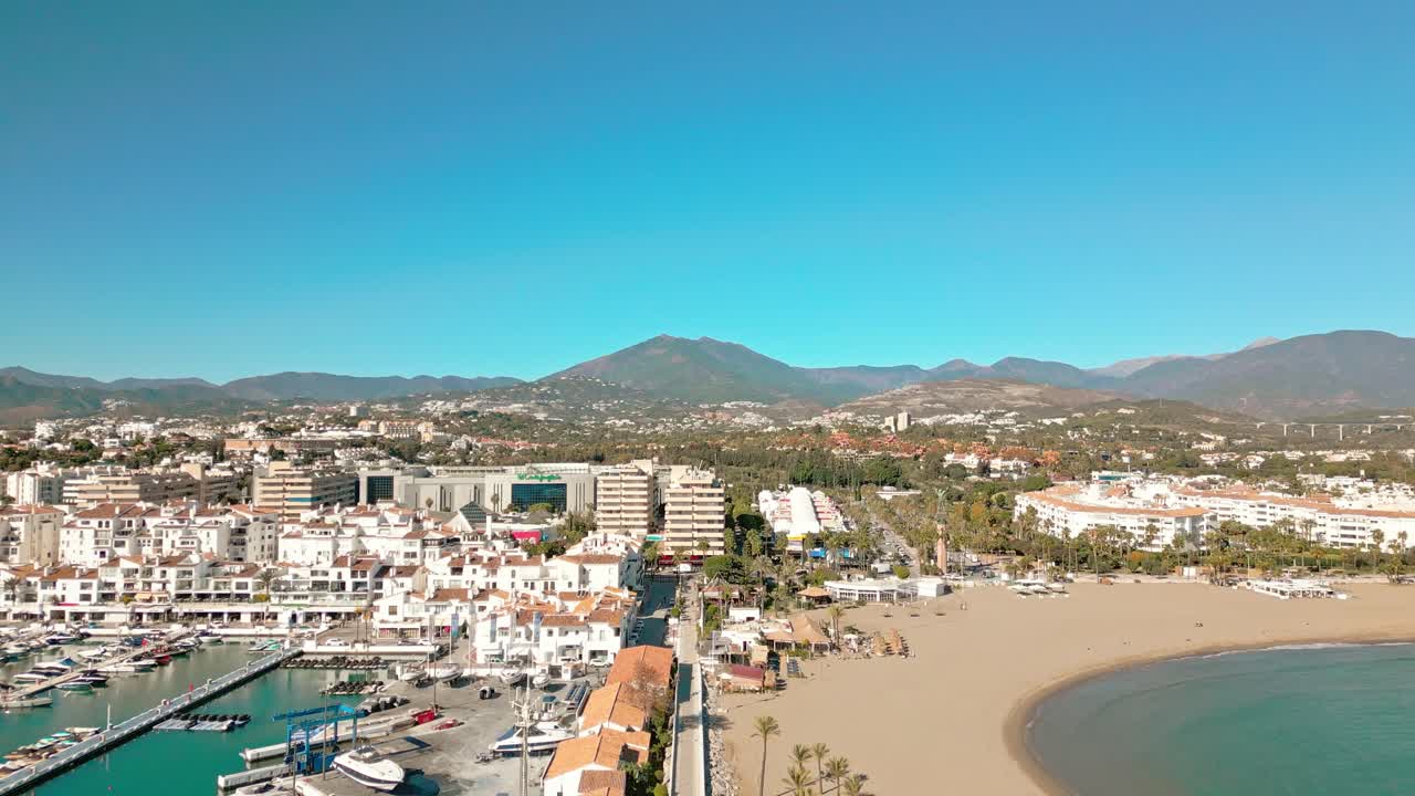 Ripresa aerea in retromarcia sopra il porto turistico di Puerto Banús e la spiaggia di Marbella, nel sud della Spagna, e le montagne La Concha sotto un cielo azzurro e soleggiato