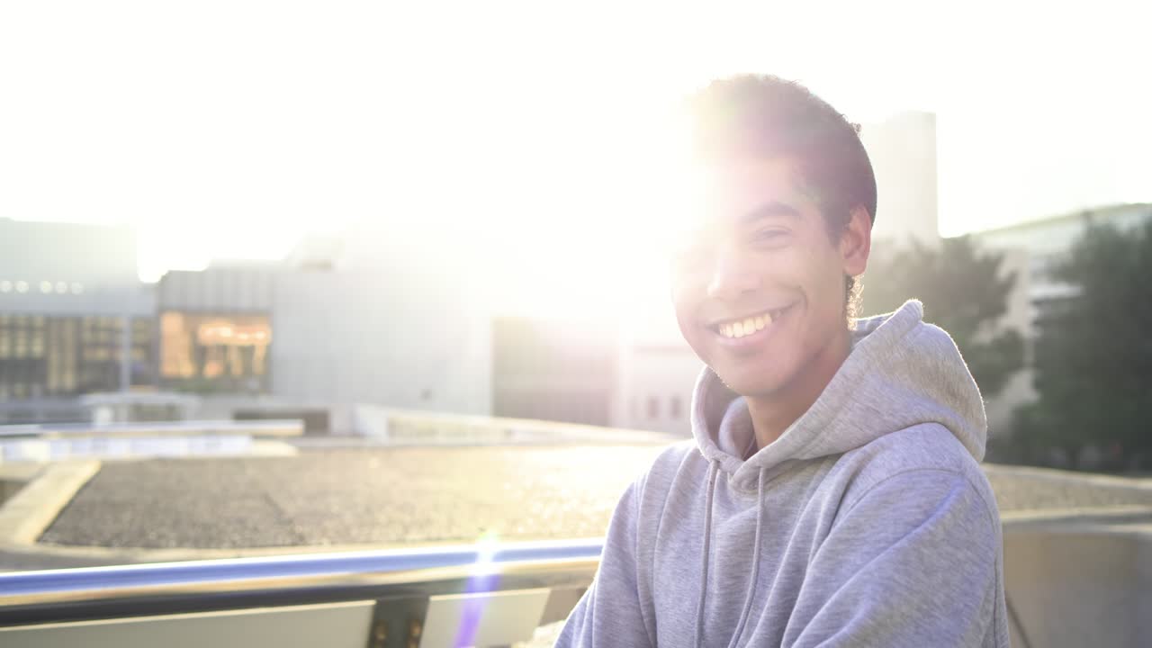 Homem afro-asiático encostado no parapeito do telhado, contraluz pelo sol, sorrindo e rindo para a câmera.