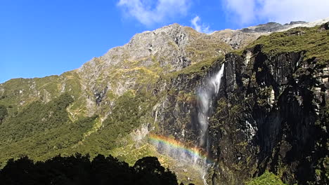 steady wide shot of the rob roy glacier's waterfall in new zealand, falling in the wind and creating a rainbow during a sunny day with blue sky