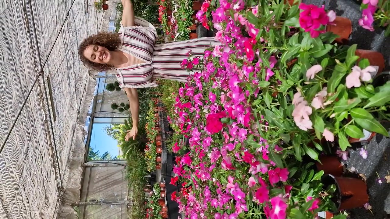 Jovem mulher feliz rodopiando com os braços para cima em um viveiro cheio de plantas floridas.