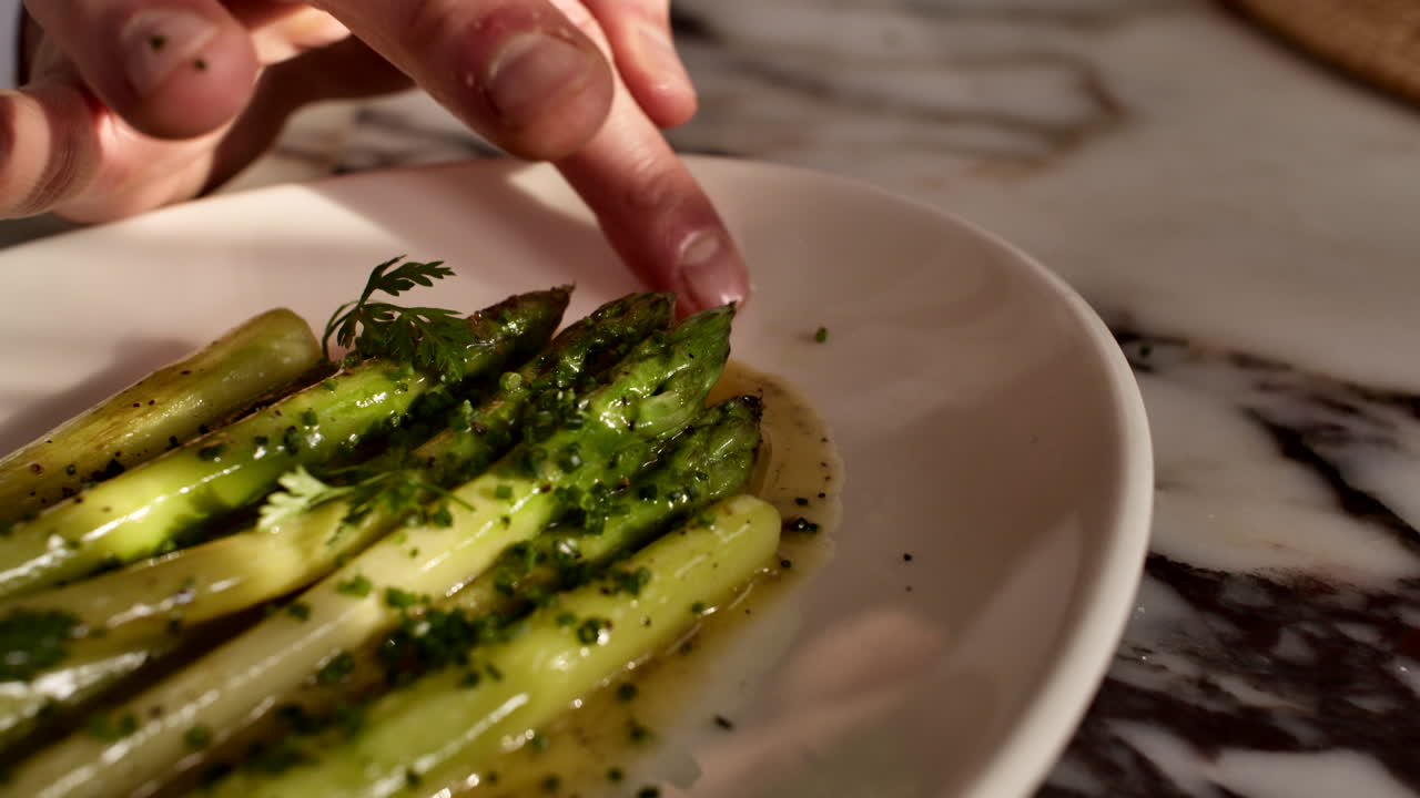 Close-up of a chef's hand adding a final herb garnish to a professional plate of seasoned asparagus in a generic kitchen. Cinematic food styling for fine dining presentation