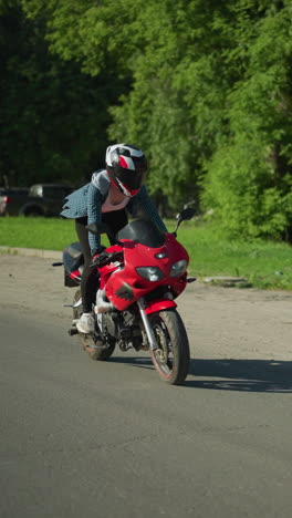 una dama monta una bicicleta eléctrica roja, de pie hábilmente mientras se equilibra en movimiento, su camisa flota en el viento, revelando parcialmente su cuerpo, navegando en un día soleado a lo largo de un camino despejado