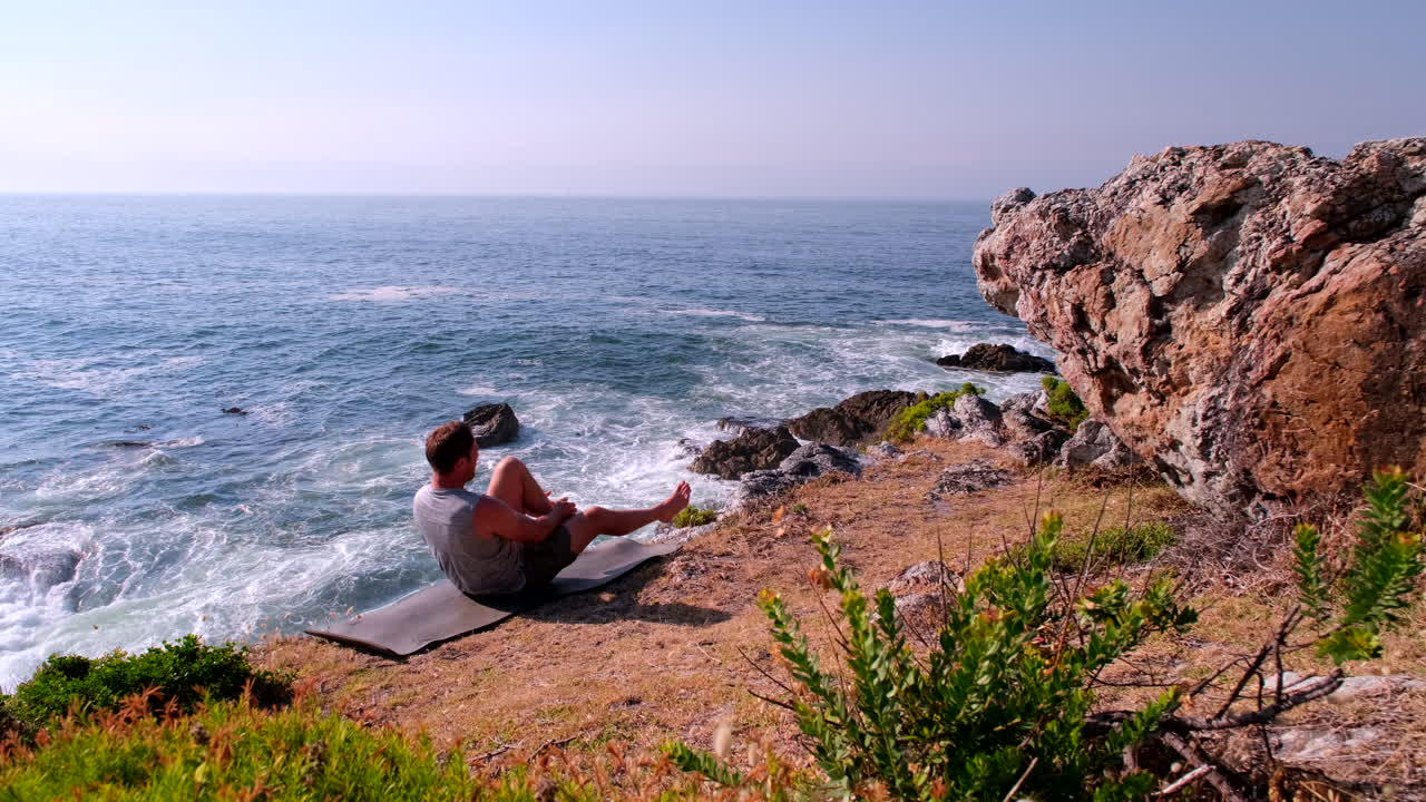 Entrenamiento abdominal con rodillas al pecho de un hombre descalzo sentado en un acantilado con vistas al mar