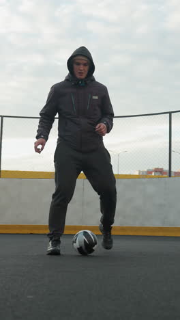atleta practicando ejercicios de fútbol en la arena deportiva, demostrando habilidades de trabajo de pies mientras controla la pelota con precisión, el fondo presenta postes de lámparas, postes de portería y edificios residenciales urbanos