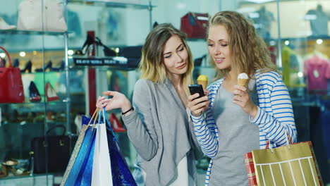 dos mujeres jóvenes con bolsas de compras dyal ir helado y considerar algo en el teléfono sonreír es