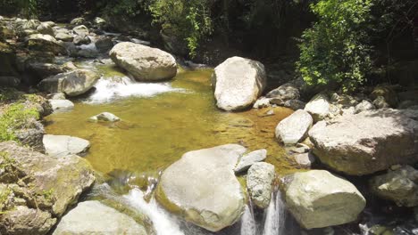 Pull-back-view-from-wooden-handrail-of-fresh-clear-mountain-stream-cascades-down-rocks-on-sunny-day,-handheld