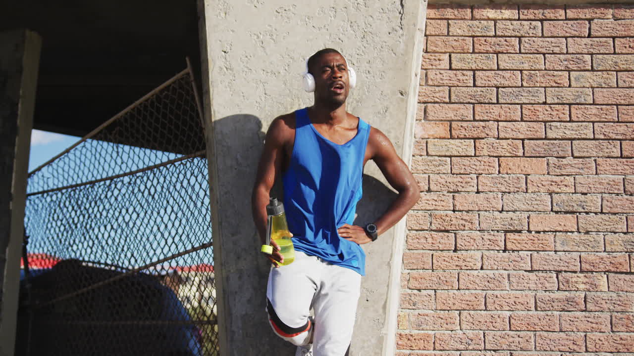 African american man wearing headphones drinking from water bottle taking break in exercise outdoors