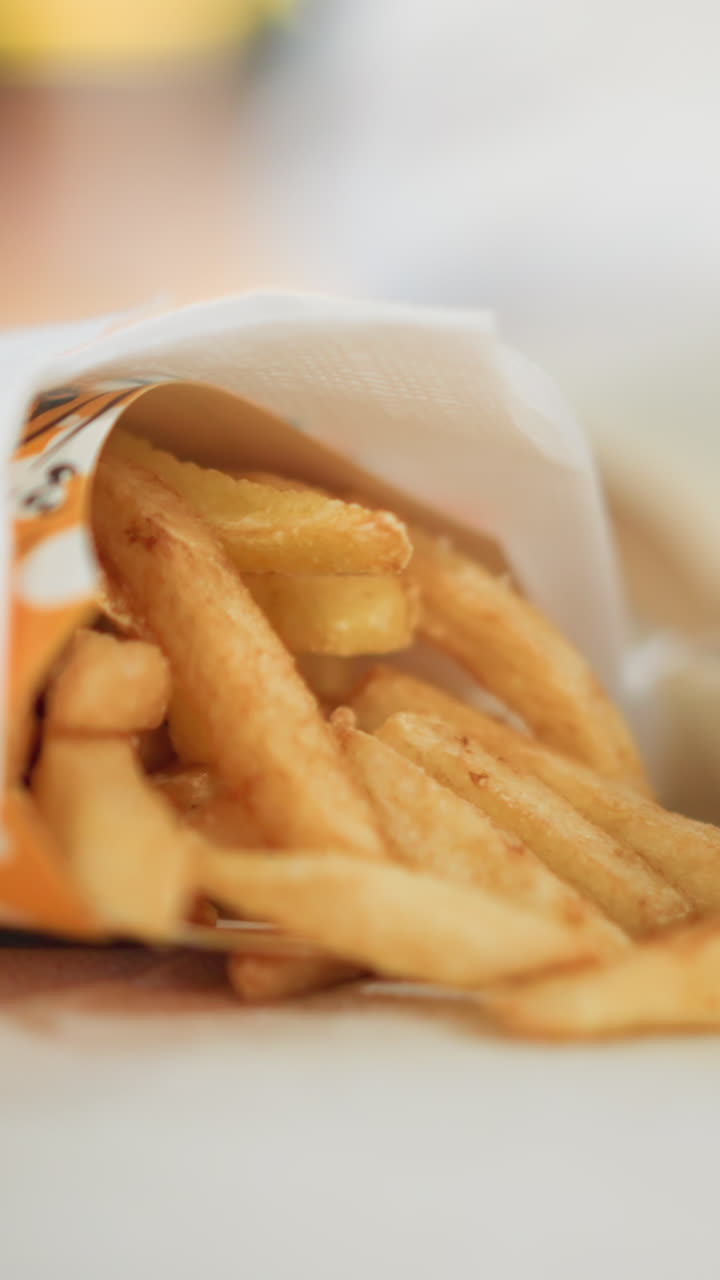 Close-up of hand reaching for crispy fries from paper bag on table in casual dining setting, soft blurred background with out-of-focus lights creates a warm