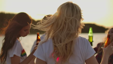 a blonde female in white t-shirt is moving her slender body and hands on the beach party on the sand river coast with beer.