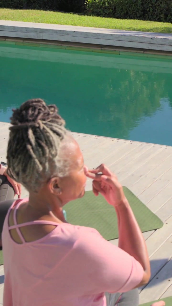 Vertical video: Pinching nostril senior in pink breathing and stretching on deck with mats for yoga