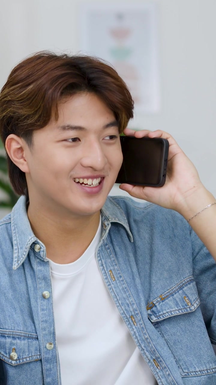 Un giovane uomo asiatico allegro con una camicia di jeans parla al suo telefono cellulare al chiuso.