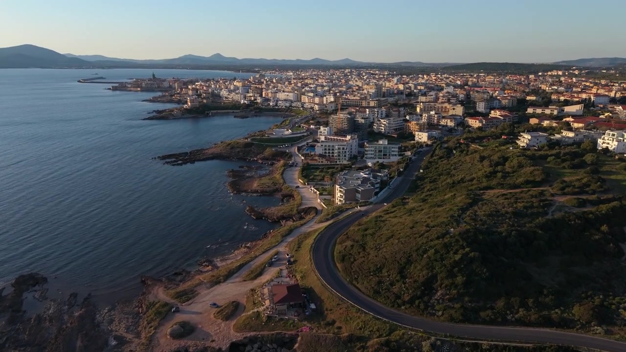 Una strada costiera tortuosa conduce verso la città di mare di Alghero mentre un drone sorvola la costa rocciosa all'ora d'oro